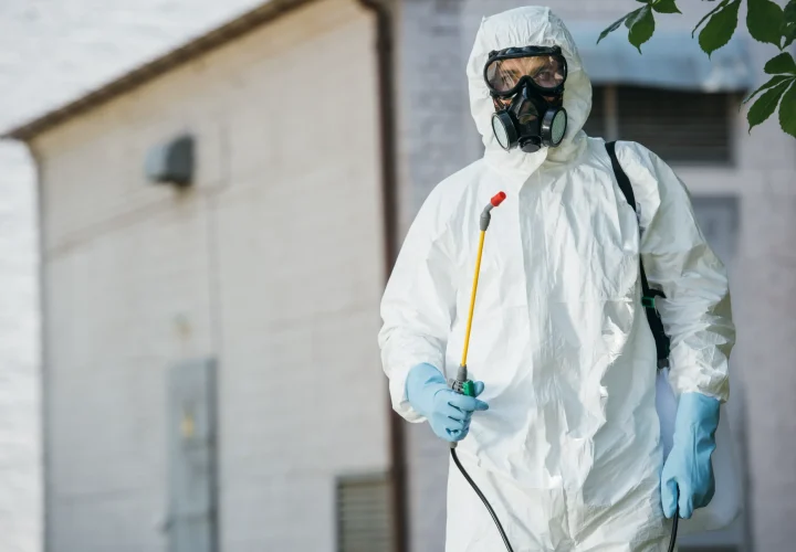 Person in protective suit and gas mask holding a sprayer stands outside a white building, suggesting focus on safety and sanitation efforts.